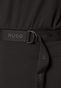 Schwarzes Kleid mit glatter Textur, ausgestattet mit einem tonalen Gurt mit einer kreisförmigen Schnalle und "HUGO"-Prägung am Riemen. Einfaches und elegantes Design.
