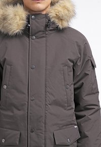 Parka grigio scuro con cappuccio in pelliccia sintetica, bottoni a pressione, due tasche frontali e una texture liscia. Presenta una tasca sulla manica in stile utility.