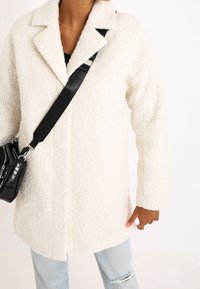 Weißer, strukturiert wirkender Kunstpelzmantel mit Reverskragen, langen Ärmeln und lässiger Passform, kombiniert mit einer schwarzen Umhängetasche und hellblauen Jeans.