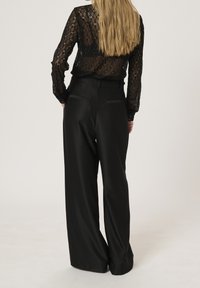 Top lungo in pizzo nero con maniche lunghe e motivi intricati abbinato a pantaloni ampi in satin nero a vita alta con tasche posteriori.