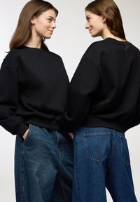 Due donne che si fronteggiano, indossando felpe nere e jeans blu, in piedi contro uno sfondo semplice e chiaro.