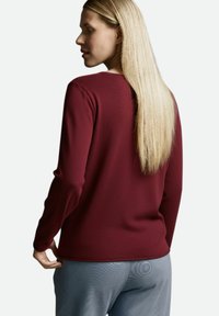 Burgunderrotes Langarmshirt aus strukturiertem Stoff mit rundem Ausschnitt und körperbetontem Schnitt, kombiniert mit hellgrauen Hosen mit dezentem Muster.