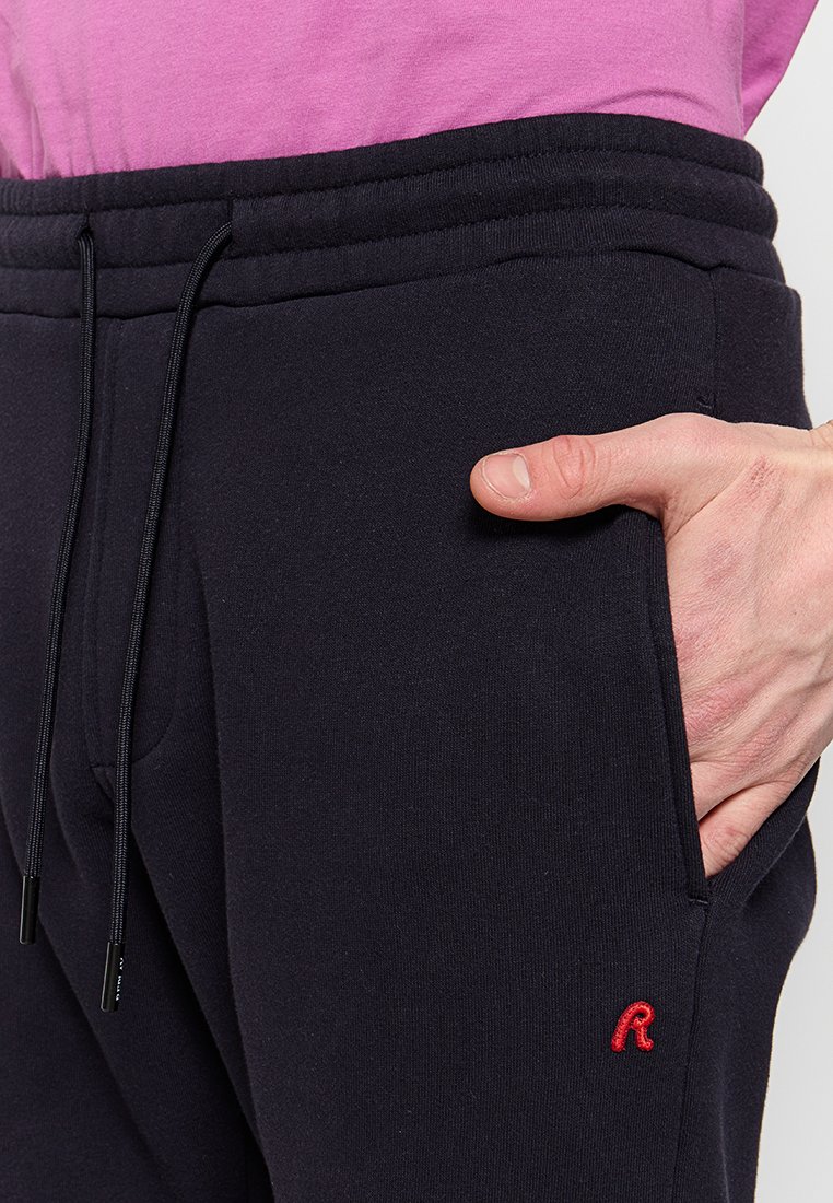 Pantaloni felpati blu navy con vita elastica e cordoncino. Dotati di tasche laterali e un piccolo logo rosso ricamato sulla coscia sinistra.