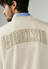 Beige sweatshirt med stor, tekstureret "BRKLYN" broderi og "Williamsburg" i skrå skrift, med ribbet krave og blød tekstur.