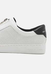 Sneaker in pelle bianca con design perforato, accentuazione nera sul tallone, zip argentata e logo Michael Kors sul lato. Suola a profilo basso.