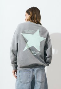 Sweat-shirt gris arborant une grande étoile vert menthe et du texte au dos, avec des poignets côtelés et une coupe décontractée. Porté avec un jean bleu clair.