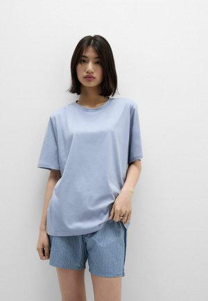 Mladá žena s tmavými vlasy po ramena má na sobě volné světle modré tričko a modré pruhované šortky, stojí před jednoduchou bílou zdí.