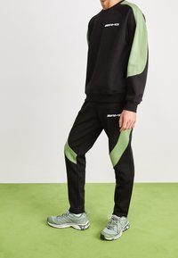 Mężczyzna ubrany w czarny i zielony dres AMG z pasującymi zielonymi butami sportowymi stoi na zielonym dywaniku.