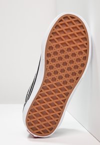 Semelle de chaussure avec motif gaufré en caoutchouc marron, dessus noir et blanc, positionnée à la verticale contre une surface blanche et un angle de mur.
