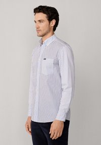 Camicia button-up a righe verticali blu e bianche. Presenta una tasca sul petto e un colletto classico. Il tessuto sembra leggero e con una texture.