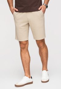 Homme portant un short beige, des baskets blanches et une chemise marron, les mains dans les poches, debout devant un fond uni.