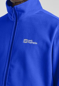 Heldblau fleecejack met hoge kraag, volledige rits en geborduurd "Jack Wolfskin" logo. Zachte textuur. Geschikt voor buitenactiviteiten.