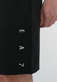 Shorts sportivi neri realizzati in materiale leggero, con un design geometrico e il logo "EA7" bianco sulla parte inferiore sinistra.