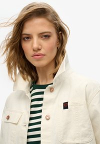 Chaqueta de lienzo blanca con un cuello alto, acentos de botones de cobre y un bolsillo de parche. Debajo, se ve una camisa de rayas verde y blanca.