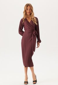 Robe portefeuille en bordeaux profond avec texture plissée. Manches longues, décolleté en V et détail de taille à nouer. Longueur genou avec une finition lisse.