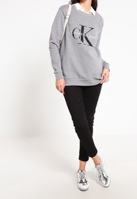 Felpa grigia con grafica nera "Calvin Klein Jeans", camicia bianca con colletto sotto, pantaloni skinny neri e sneakers argento lucido.