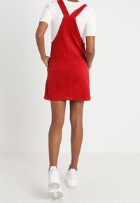 Rotes Kord-Overall-Kleid mit überkreuzten Trägern, aus einem strukturierten Stoff und in lockerer Passform, getragen über einem weißen kurzärmligen Oberteil.