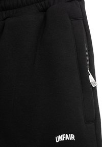 Svarte joggebukser i teksturert stoff, med elastisk midjebånd og en lomme med glidelås på siden. "UNFAIR" er brodert i hvitt.
