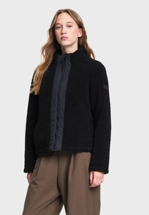 Jeune femme portant une veste polaire noire zippée avec un pantalon marron ample, debout devant un fond clair uni.