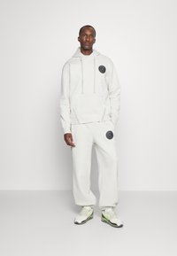 Hvid svedhætte med justerbare snoretræk og en frontlomme, parret med matchende joggers med en sort logo-patch. Struktureret stof.