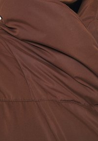 Hnědá polstrovaná tkanina s hladkým povrchem, s prošitím podél švů a jemným drapováním pro zvýšení designových detailů.