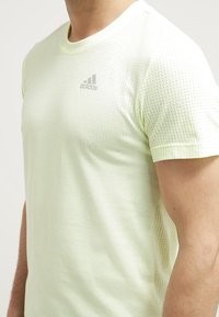 Camiseta atlética amarilla clara con un patrón de malla texturizada y mangas cortas, que presenta un sutil logo gris de Adidas en el pecho.