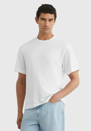 Man med kort lockigt hår som bär en enkel vit t-shirt med kort ärm och ljusblå jeans, stående med ena handen i fickan.