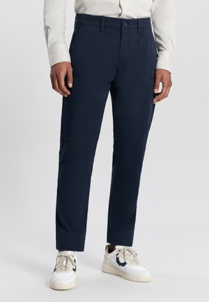 Pantalon tailleur bleu marine en tissu lisse, avec une taille plate, des poches latérales et une fermeture à bouton. Associé à des baskets blanches sportives.
