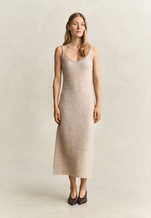 Femme aux longs cheveux blonds portant une robe sans manches, beige clair, longueur cheville, et des chaussures sombres à bout pointu, debout devant un fond uni.