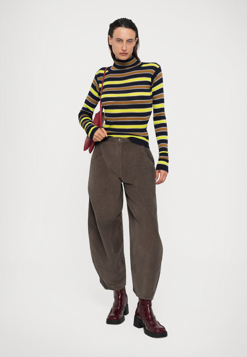 Donna che indossa un dolcevita a righe gialle, marroni e nere, pantaloni in corduroy marroni e larghi, stivaletti borgogna e porta una borsa a tracolla rossa.