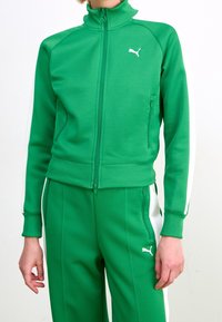 Veste de survêtement zippée verte en tissu texturé, avec un col haut et des bandes blanches sur les côtés. Logo Puma visible sur la poitrine.