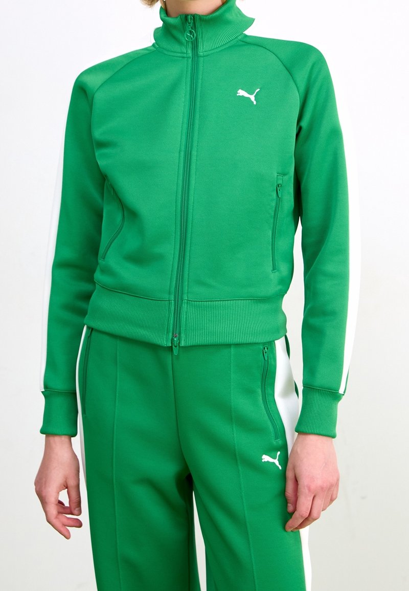 Veste de survêtement zippée verte en tissu texturé, avec un col haut et des bandes blanches sur les côtés. Logo Puma visible sur la poitrine.