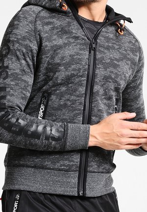 Mann trägt grauen Camo-Kapuzenpullover mit Reißverschluss, "Superdry"-Markenaufdruck am Ärmel und mit Reißverschlusstaschen, kombiniert mit schwarzen Sporthosen.