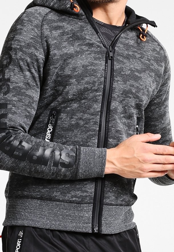 Homme portant un hoodie zippé gris camouflage avec le logo "Superdry" sur la manche et des poches zippées, assorti à un pantalon de sport noir.