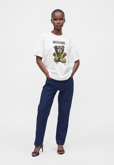 Άτομο που φοράει λευκό oversized t-shirt Moschino με γραφικό αρκουδάκι και κίτρινη ταινία, σκούρο μπλε τζιν και μαύρα σανδάλια με τακούνι.