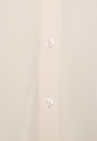 Blouse rose pâle en tissu lisse, avec fermeture classique à boutons à quatre trous et coutures discrètes le long de la patte de boutonnage.