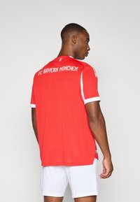 Rød FC Bayern München-drakt med hvite detaljer, korte ermer og klubbnavn trykt i hvitt på baksiden. Lett stoff med et klassisk design.