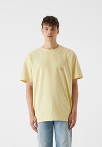 Geltonas medvilninis marškinėliai su laisvu kirpimu, trumpomis rankovėmis ir maža grafiką švelnią spalva ant krūtinės. Suderinti su šviesiai mėlynomis džinsais.