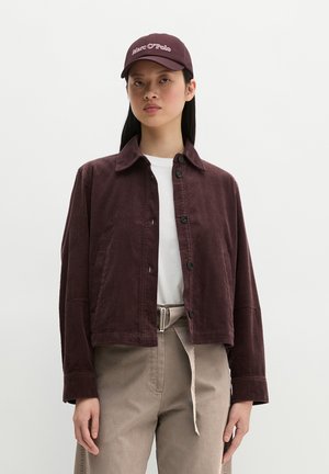 Femme portant une veste en velours côtelé violet foncé, un t-shirt blanc, un pantalon beige avec ceinture et une casquette violet foncé avec texte.
