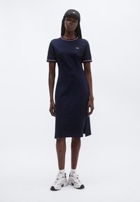 Γυναίκα με μπλε μεσάτου μήκους φόρεμα με κοντά μανίκια, άσπρα αθλητικά παπούτσια και μαύρο καπέλο, στέκεται μπροστά σε λευκό απλό φόντο.