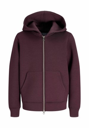 Garçon blond regardant vers la droite, portant un sweat à capuche zippé bordeaux, un t-shirt blanc, un pantalon beige large et des chaussures beiges sur un fond clair uni.