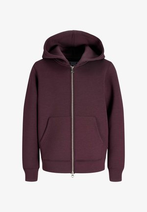 Garçon blond regardant vers la droite, portant un sweat à capuche zippé bordeaux, un t-shirt blanc, un pantalon beige large et des chaussures beiges sur un fond clair uni.