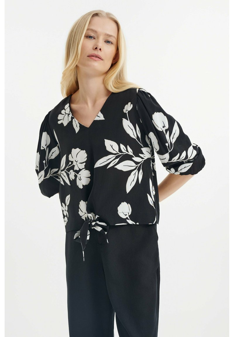 Schwarze Bluse mit weißem Blumenmuster, V-Ausschnitt, Puffärmeln und Bindeband in der Taille. Weicher Stoff mit fließendem Design.