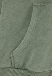Gros plan sur un tissu vert montrant une poche avec des coutures et un ourlet côtelé, probablement une partie d’un sweat-shirt ou d’un hoodie.