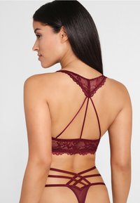 Burgunderfarbener Spitzen-Bralette mit gekreuzten Rückenriemen, der florale Muster und Netzdetails aufweist, eine weiche Textur hat und verstellbar ist.