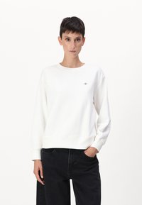 Personne aux cheveux courts et foncés portant un sweat-shirt blanc uni et un pantalon noir, debout avec une main dans la poche, sur un fond blanc.