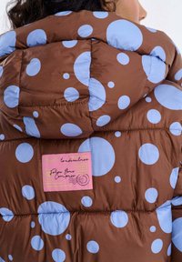Brązowa pikowana kurtka z dużymi niebieskimi kółkami, różową metką z napisem oraz kapturem, podkreślająca pikowaną fakturę i zabawny design.