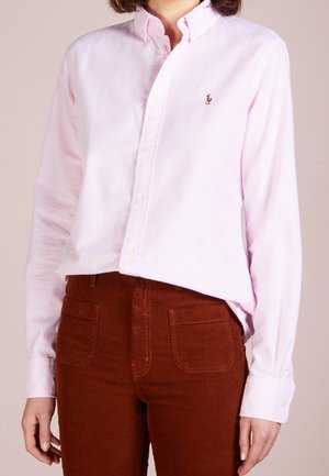Vrouw draagt een lichtroze overhemd met knopen en een klein geborduurd logo, en roestkleurige corduroy broek.