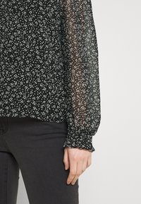 Svart genomskinlig blus med vita spridda prickar, med långa ärmar och samlade manschetter. Kombinerad med mörkgrå jeans.