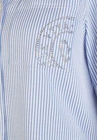 Blau-weiß gestreiftes Hemd mit einer Brusttasche, versehen mit einem silbernen Strassdesign. Baumwollstoff mit glatter Textur.